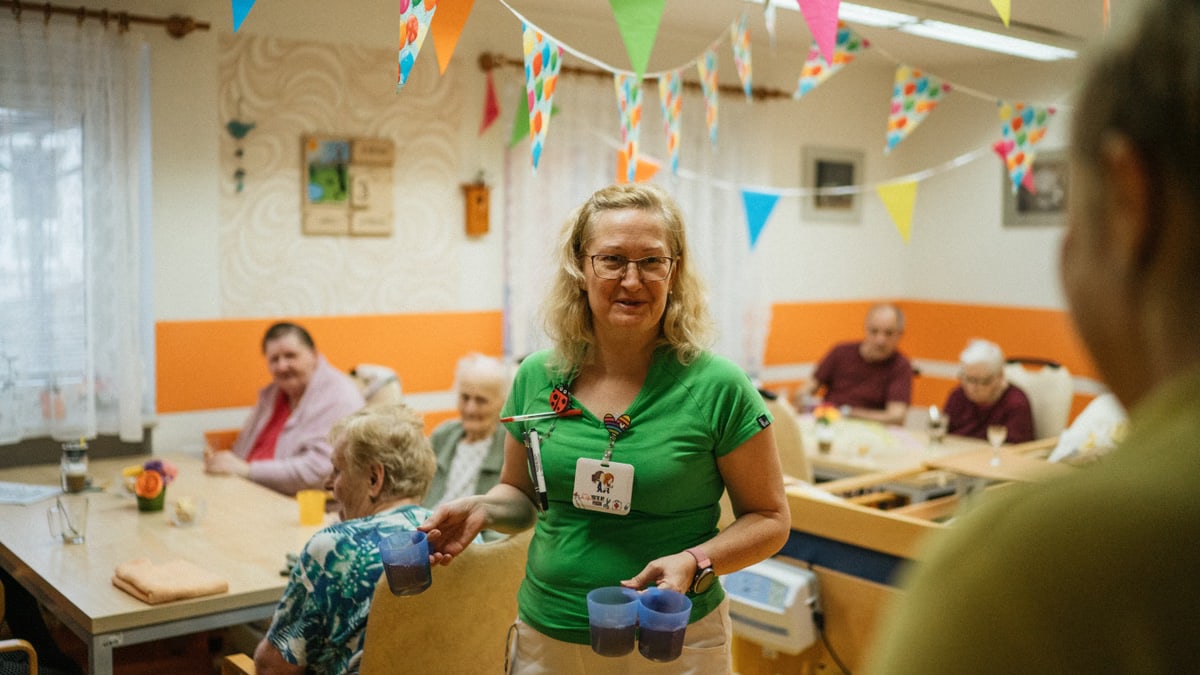 A caregiver serving drinks to elderly residents in a cheerful nursing home setting.